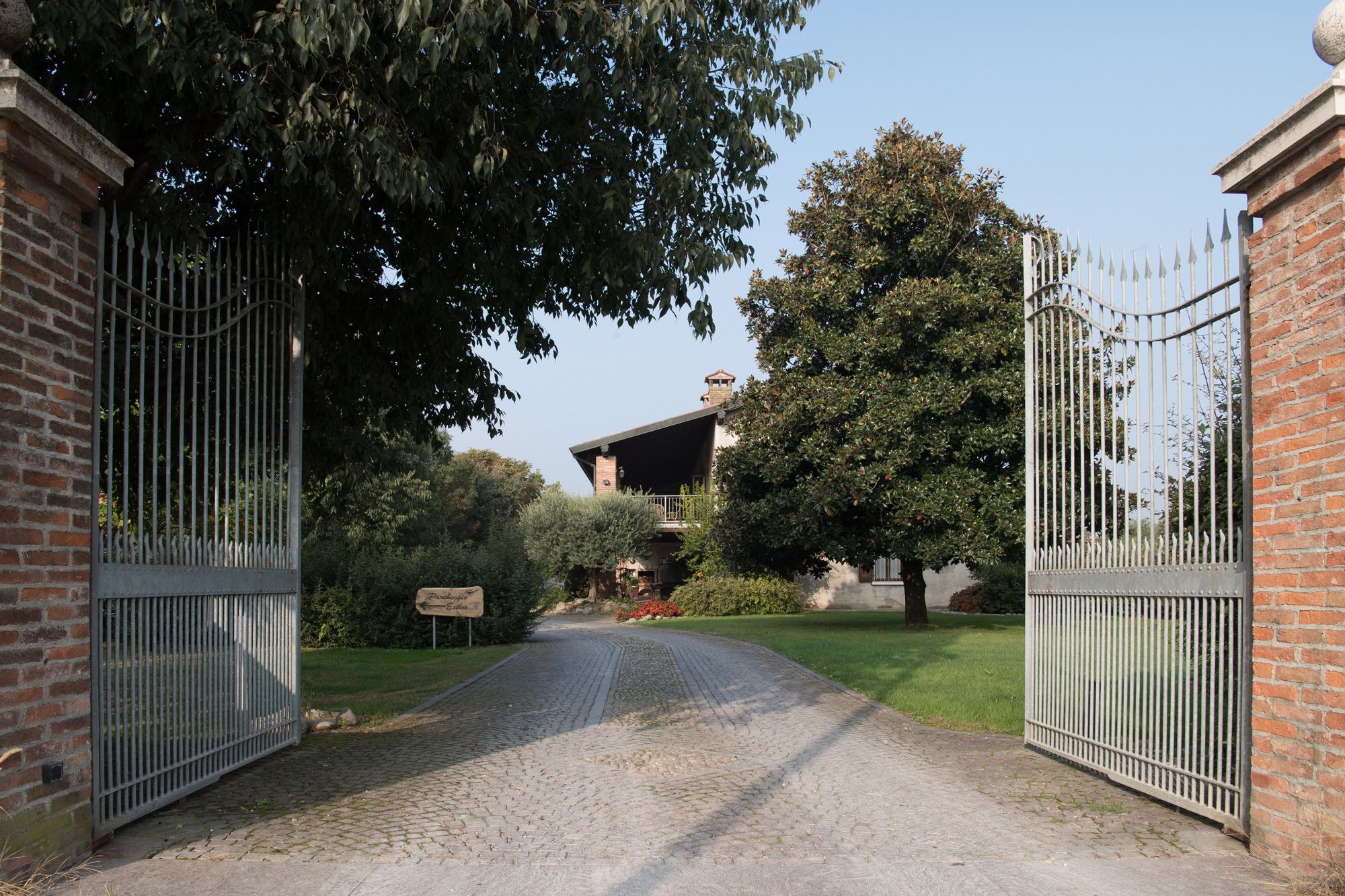 Un camino de adoquines conduce a una entrada con puerta y un edificio de ladrillo, con un gran árbol al fondo.