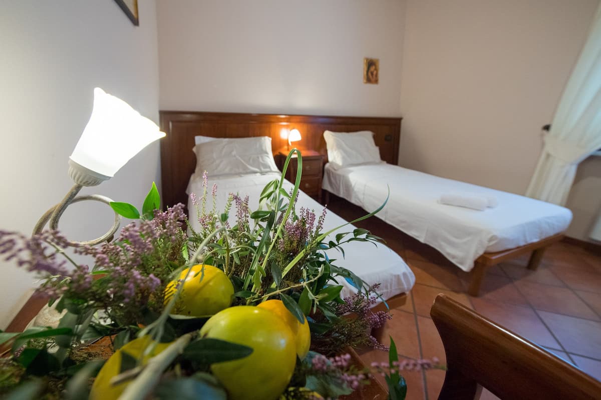 A hotel room with two single beds, a bedside table with a lamp, and a floral arrangement on the table.