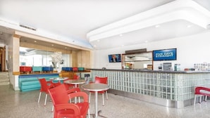 A modern hotel lobby with a bar area, seating with red chairs, and a television screen displaying 'FIVE DAYS STRAIGHT'.