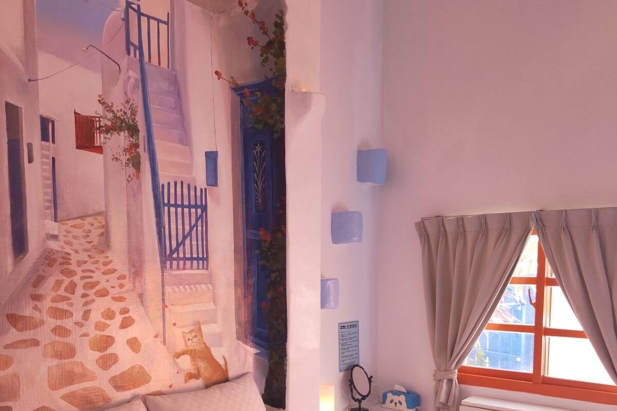Un dormitori amb un llit, una finestra amb cortines, un radiador, una taula petita amb un mirall i una paret amb un quadre.