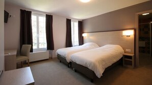 Habitación de hotel con dos camas, un escritorio y una silla. Cuenta con una ventana con cortinas, un radiador y una puerta que da a otra habitación.