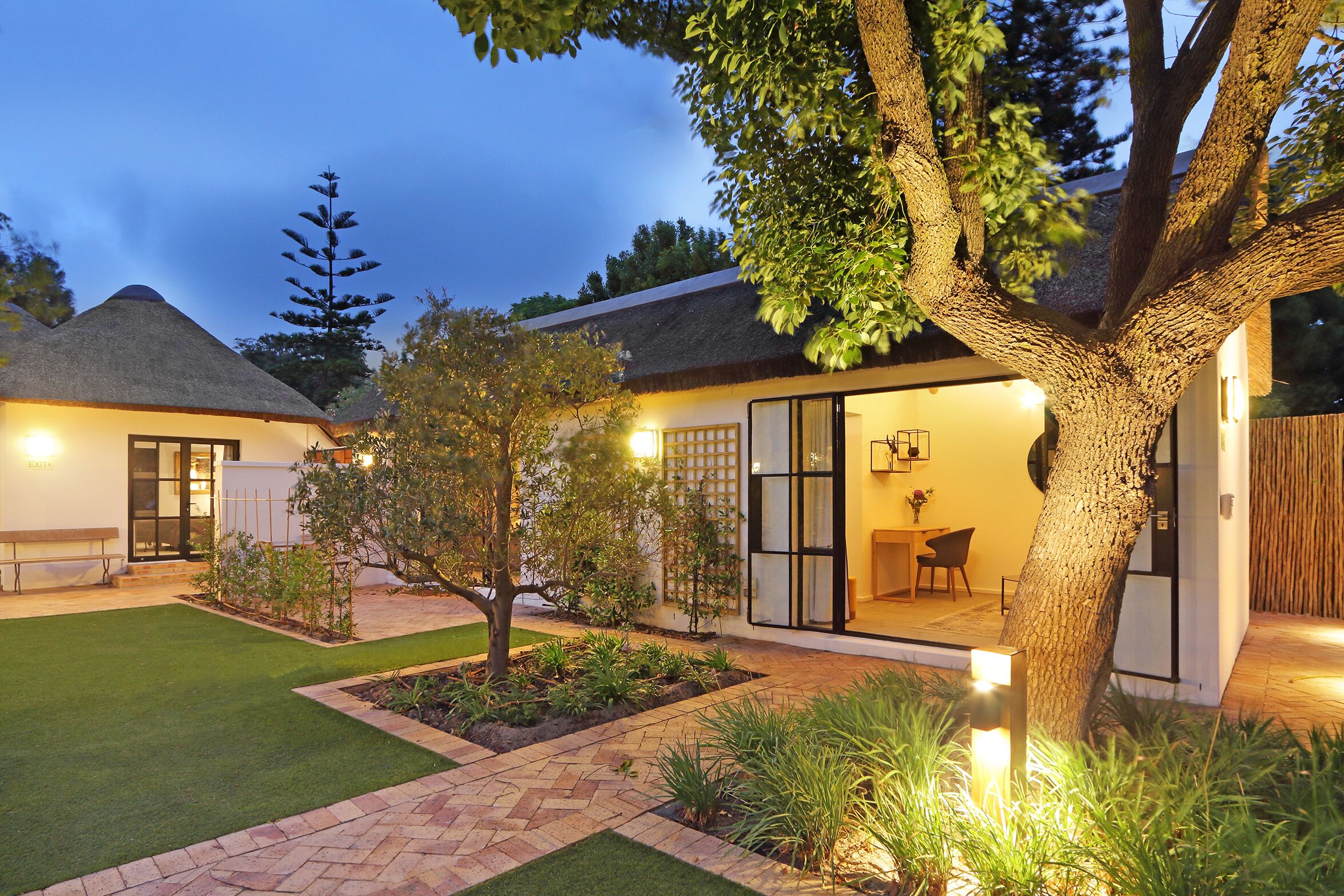 A well-lit residential property with a thatched-roof house, a paved patio, and a landscaped garden.