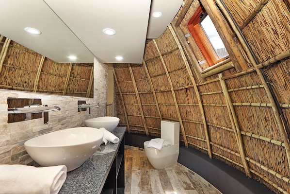 Et moderne badeværelse med et stort badekar, toilet og en vaskebord med to håndvaske. Væggene er beklædt med et bambuslignende materiale, og der er et ovenlys over toilettet.