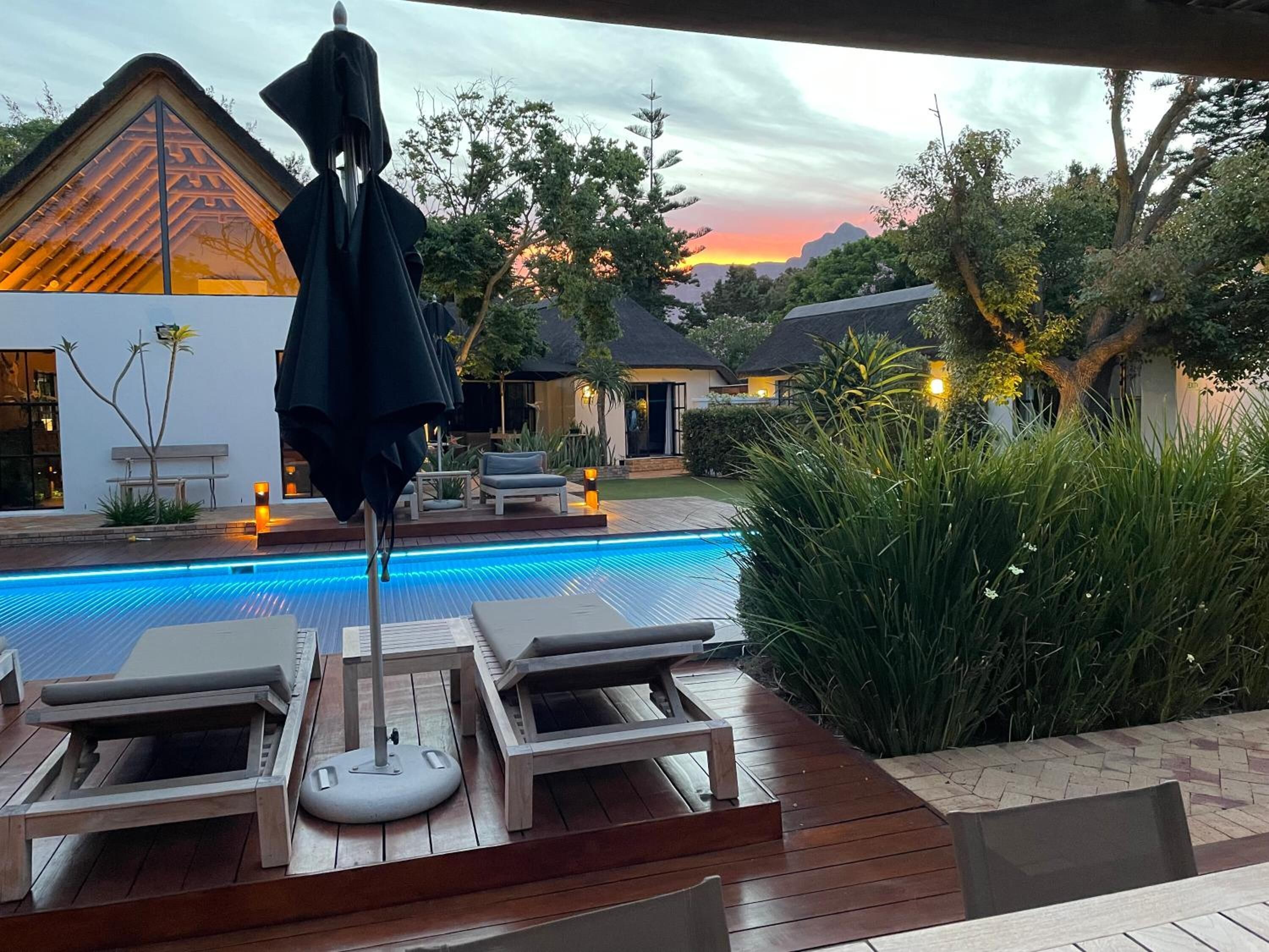 A poolside area with lounge chairs and a table, surrounded by greenery and a view of a sunset.