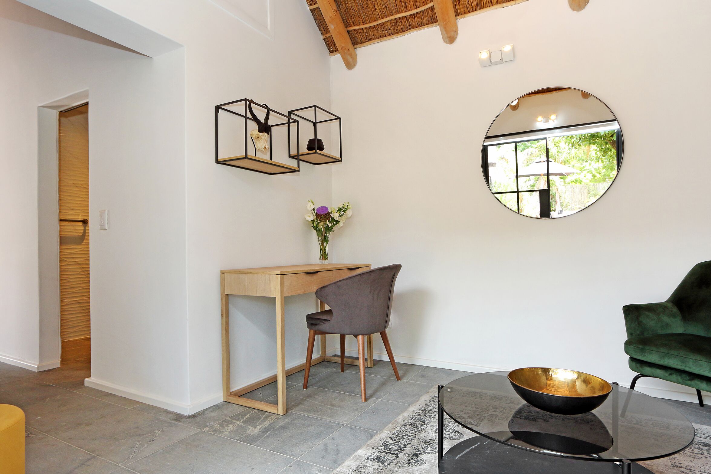 A modern room with a wooden desk, a green chair, a round mirror, and a glass coffee table with a bowl.