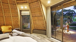 Una habitación con techo curvo, una cama con ropa blanca, una silla amarilla y un ventanal de vidrio que da a un espacio exterior con mesa y sillas.