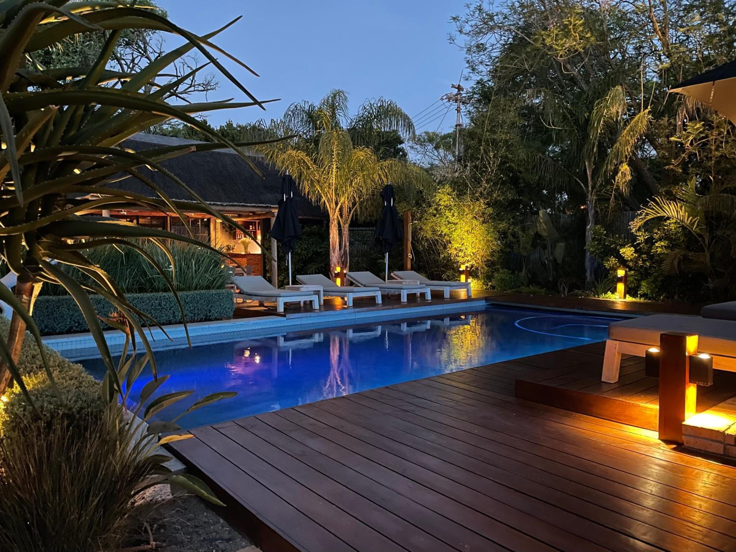 A swimming pool with lounge chairs and a wooden deck, surrounded by tropical plants and a building with a thatched roof.