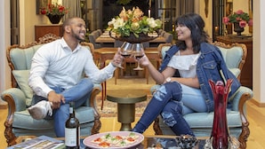 Un hombre y una mujer sentados en un sofá, sosteniendo copas de vino.  Hay una botella de vino, un plato con comida y un jarrón con flores sobre una mesa que los separa.