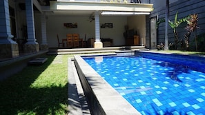 A swimming pool with blue tiles, surrounded by a grassy area and a building with a covered patio. - Krisna Seminyak (Seminyak)