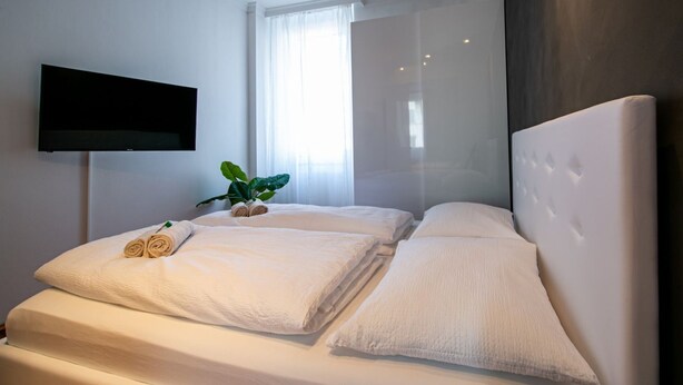 Une chambre d’hôtel moderne avec un grand lit, du linge de lit blanc, un téléviseur à écran plat fixé au mur et une grande fenêtre avec des rideaux légers.