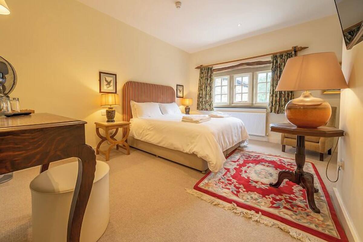 A bedroom with a bed, bedside tables, a desk, a chair, a window with curtains, and a lamp.