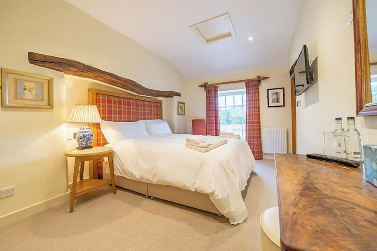 A bedroom with a bed, a bedside table with a lamp, a wooden headboard, a window with curtains, and a wooden dresser.