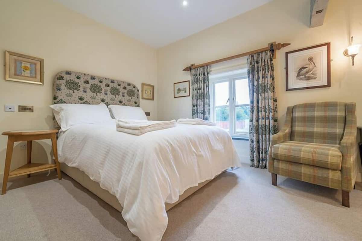A bedroom with a large bed, a wooden nightstand, a plaid armchair, and a window with curtains.