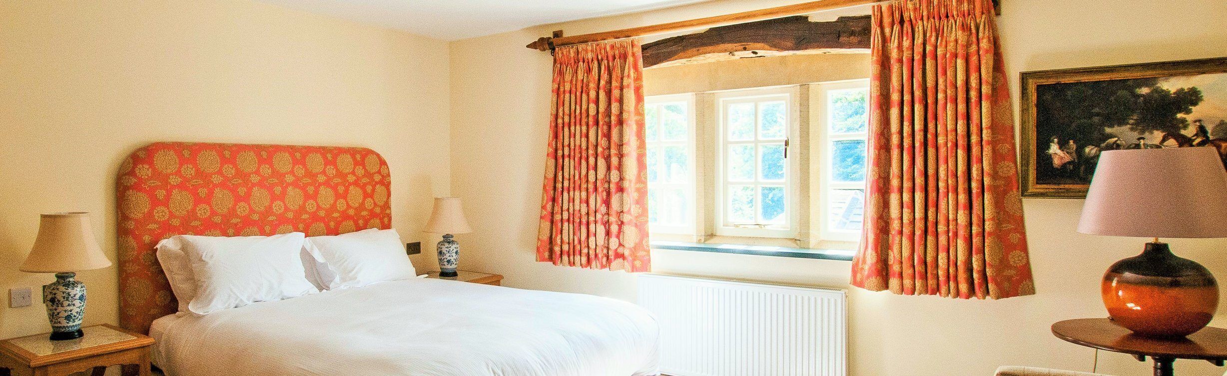 A hotel room with a bed, a nightstand, a lamp, a window with curtains, and a painting on the wall.