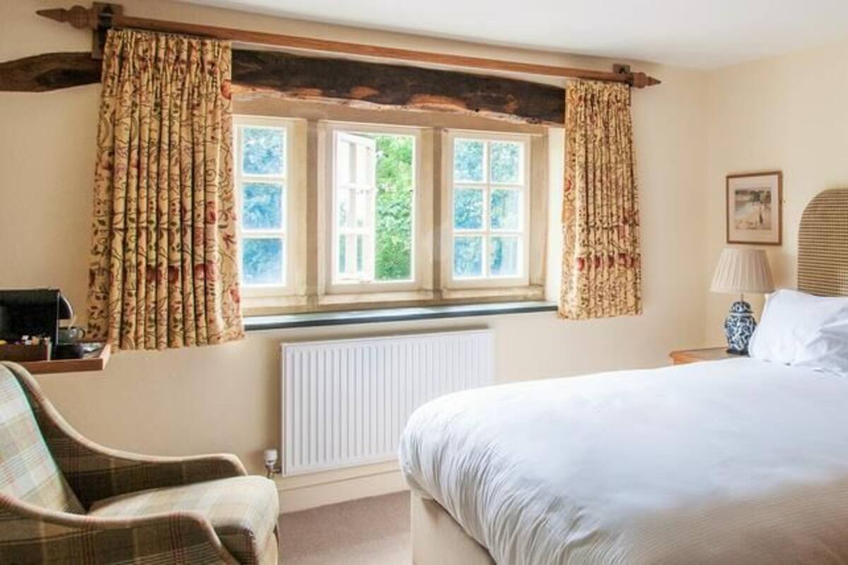 A bedroom with a large bed, a chair, a window with curtains, a radiator, and a lamp on a bedside table.