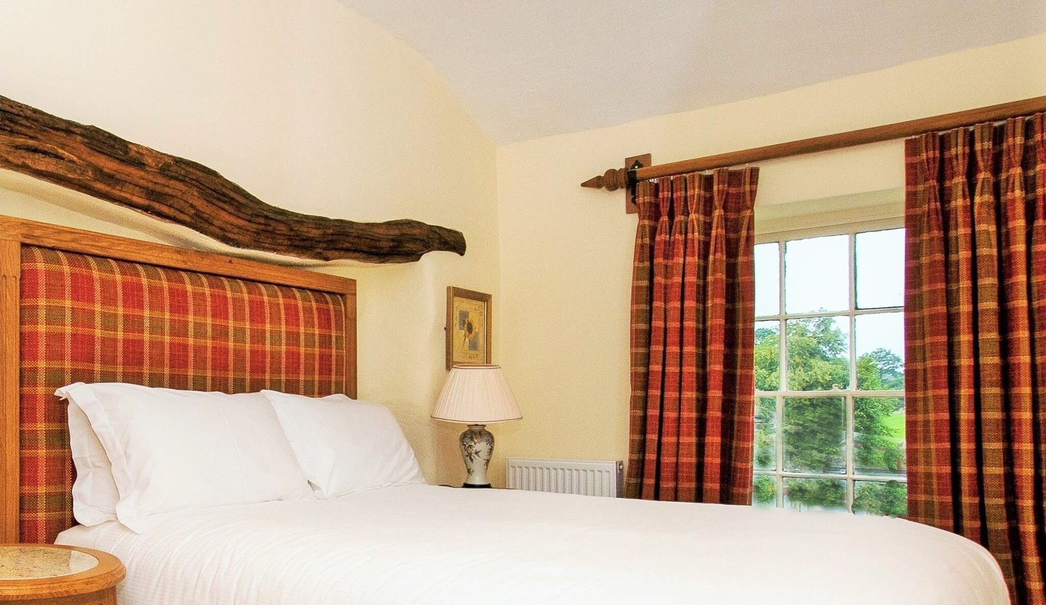 A bedroom with a large bed, plaid curtains, a wooden headboard, a bedside table, and a window with a view of greenery.