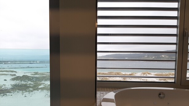 Habitación con una ventana grande que da a una playa y al océano, una bañera y suelo de baldosas.