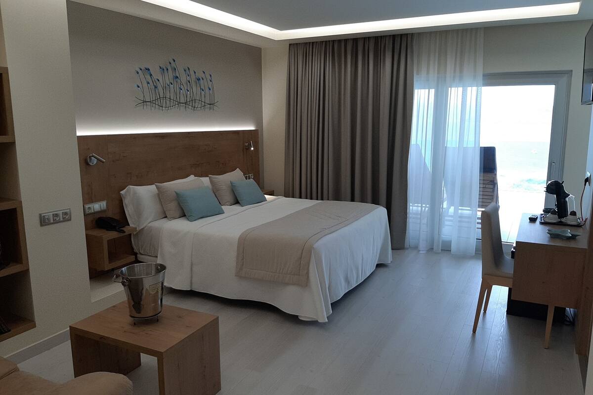 A modern hotel room with a large bed, a desk, a chair, a bedside table, and a view of the sea.