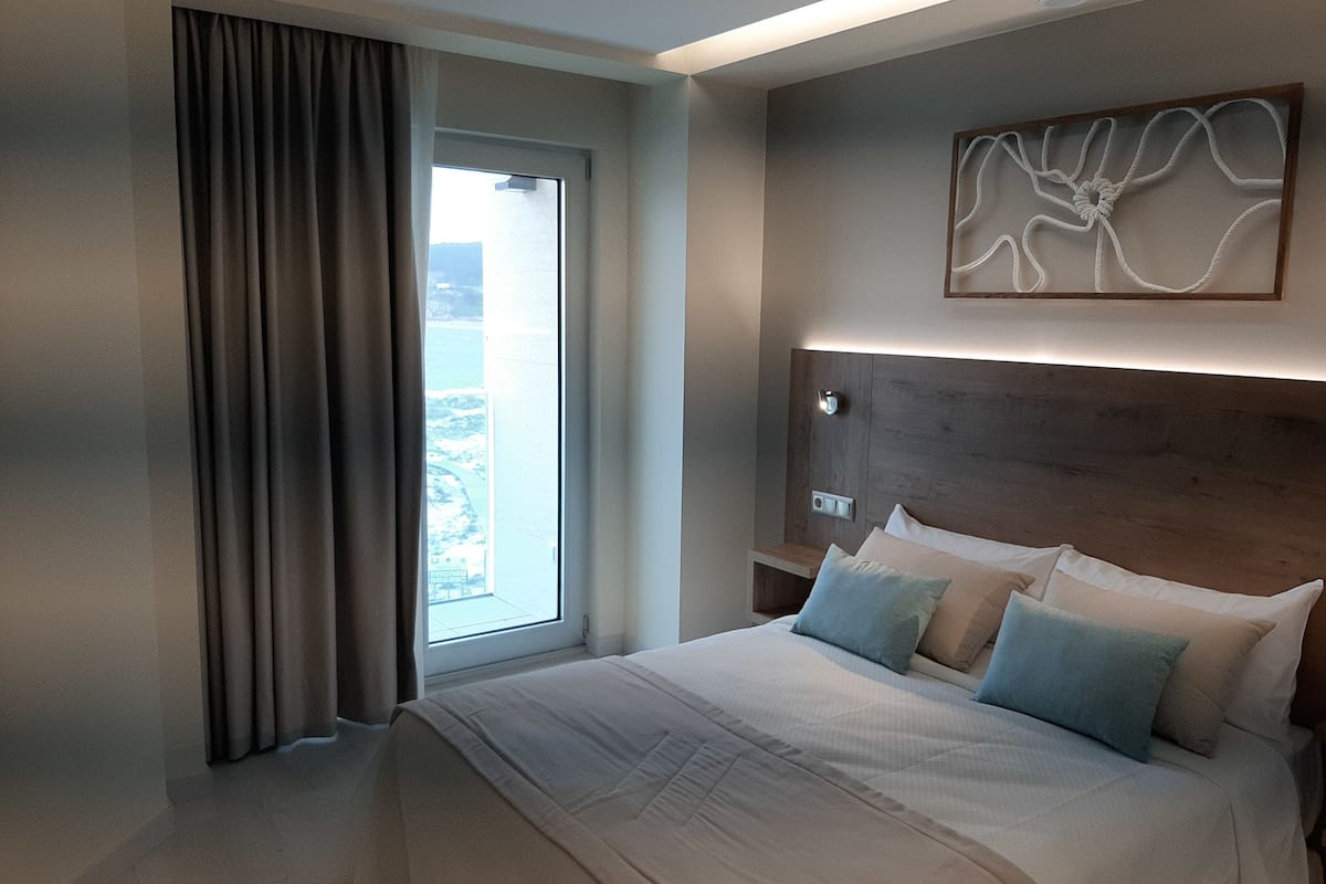 A modern hotel room with a large bed, a view of the outdoors through a window, and a wooden headboard with a light fixture.