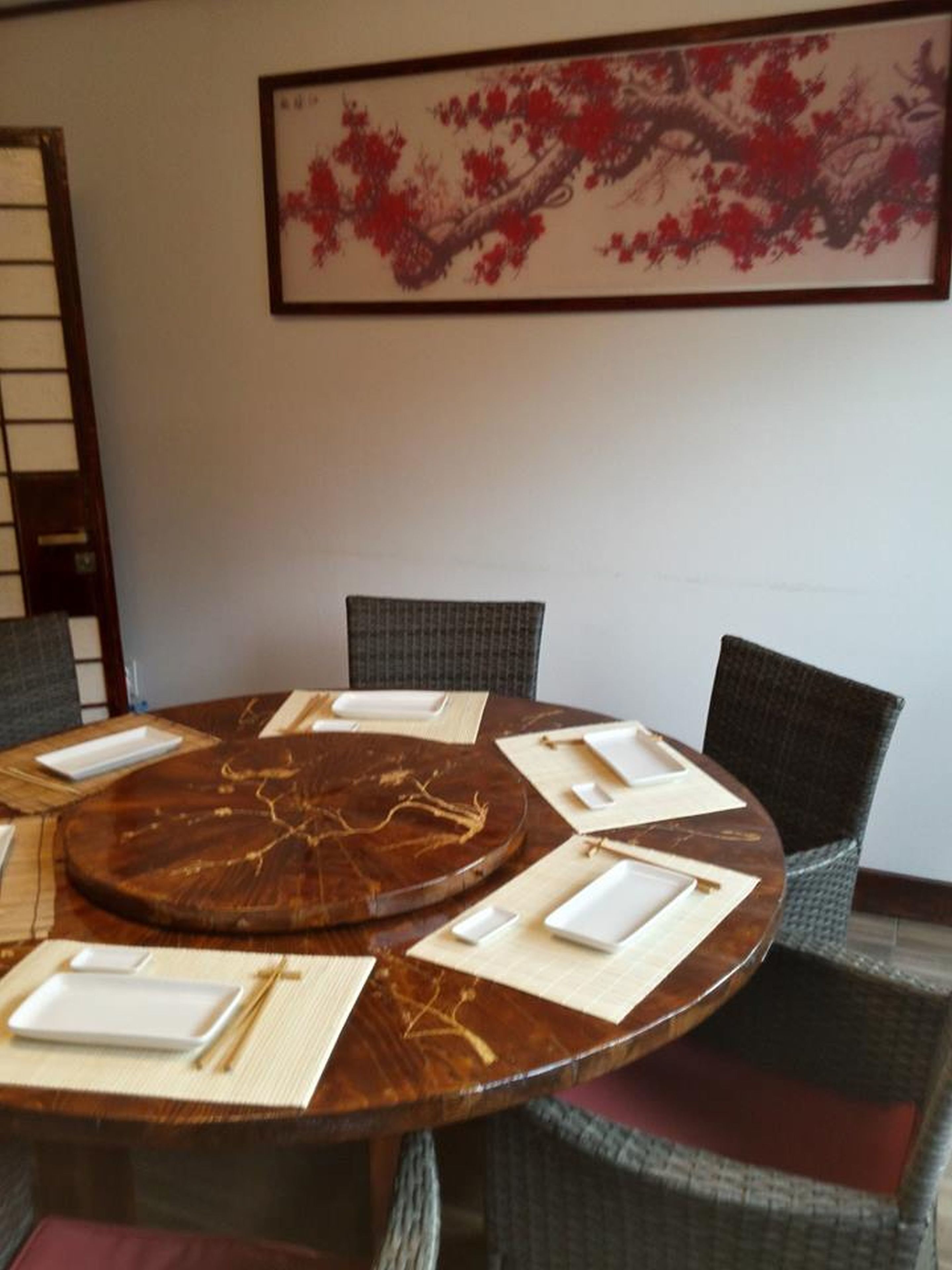 Una mesa redonda para comer con individuales blancos y palillos, rodeada de sillas de mimbre.