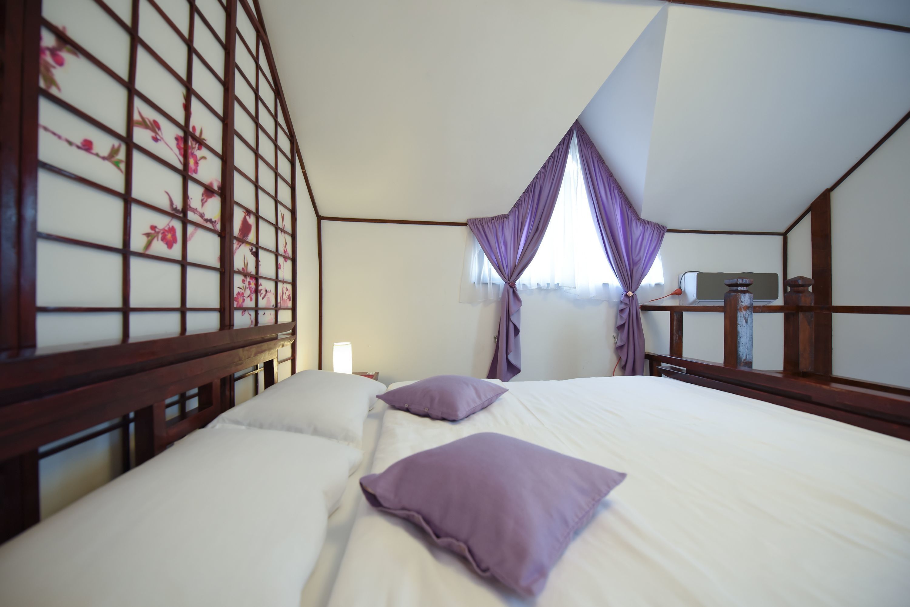 Un dormitorio con cabecera de madera, cama con ropa blanca y almohadas moradas, mesita de noche con lámpara y ventana con cortinas moradas.