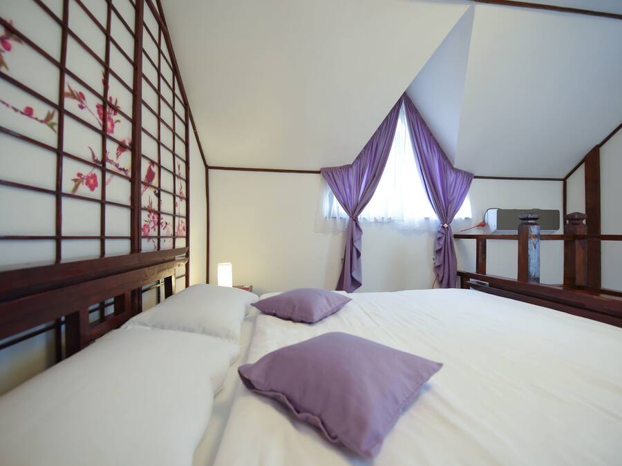 A bedroom with a wooden headboard, a bed with white linens and purple pillows, a nightstand with a lamp, and a window with purple curtains.