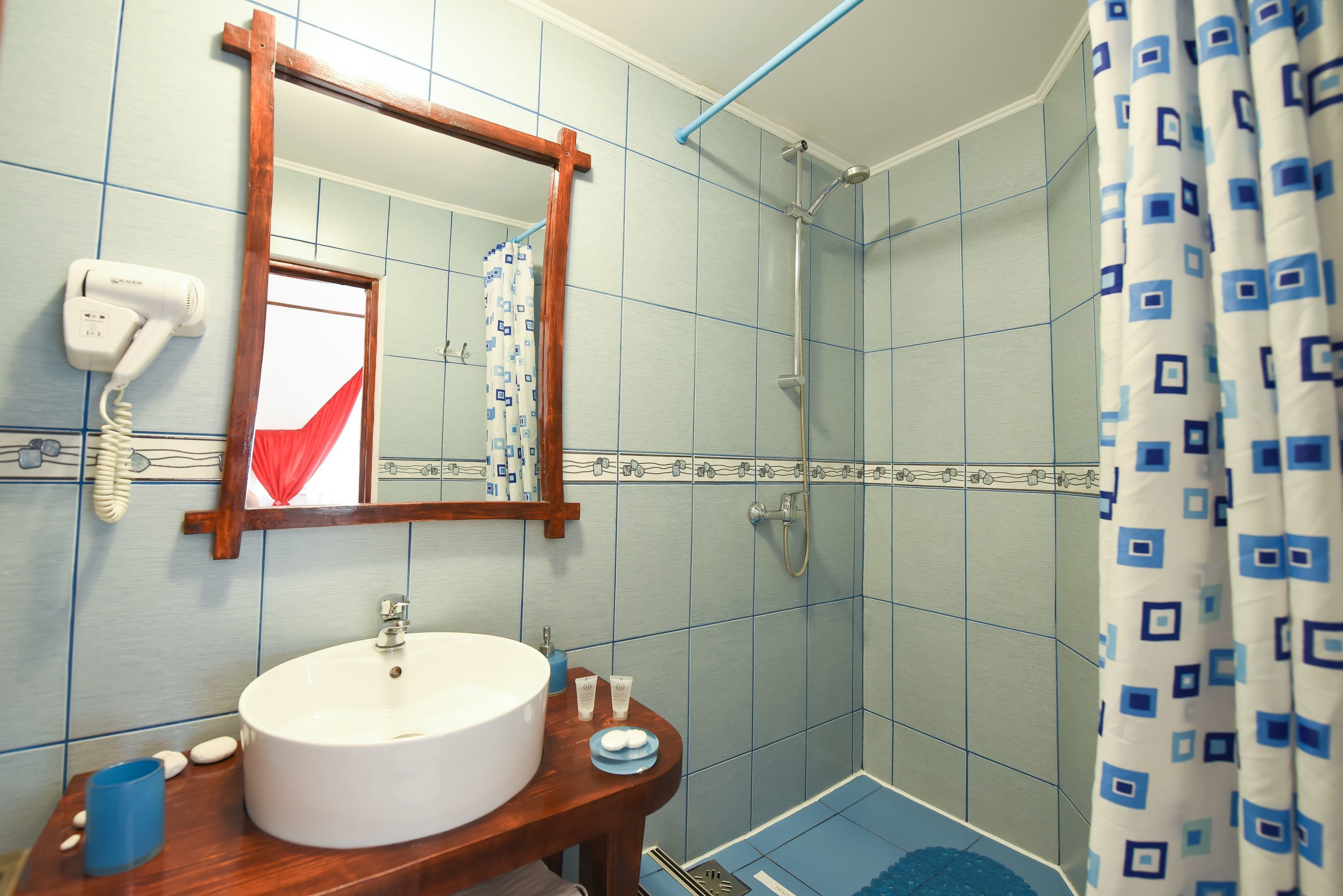 Une salle de bain avec un lavabo blanc, un meuble-lavabo en bois et une douche aux carreaux bleus et dotée d’un rideau à motifs.