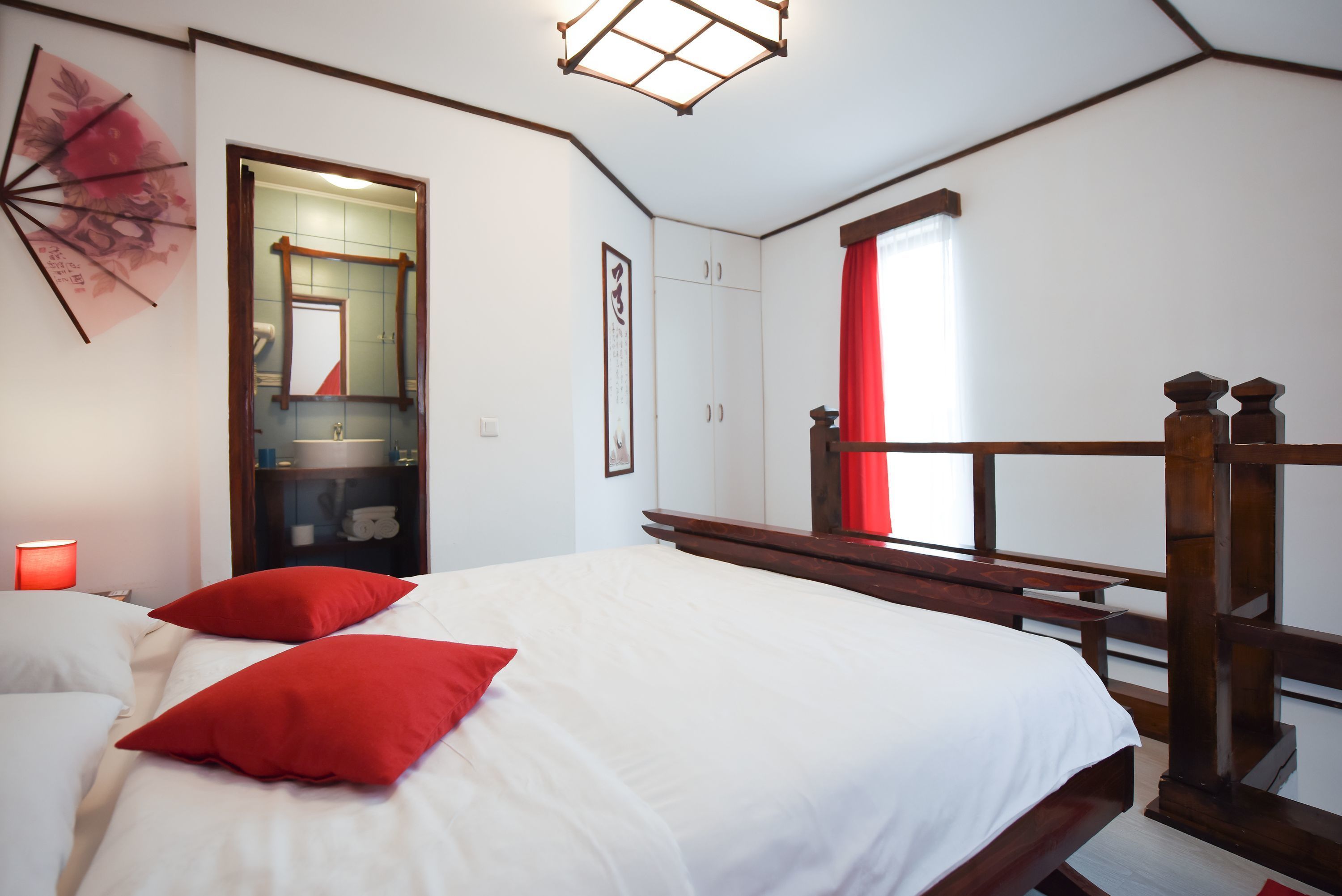 A bedroom with a bed, red pillows, a wooden headboard, a nightstand, a mirror, and a window with red curtains.