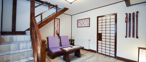 Une pièce de style japonais traditionnel, avec un sol recouvert de tatamis, une basse table en bois, un canapé violet, une porte coulissante de type shoji et un escalier en bois.