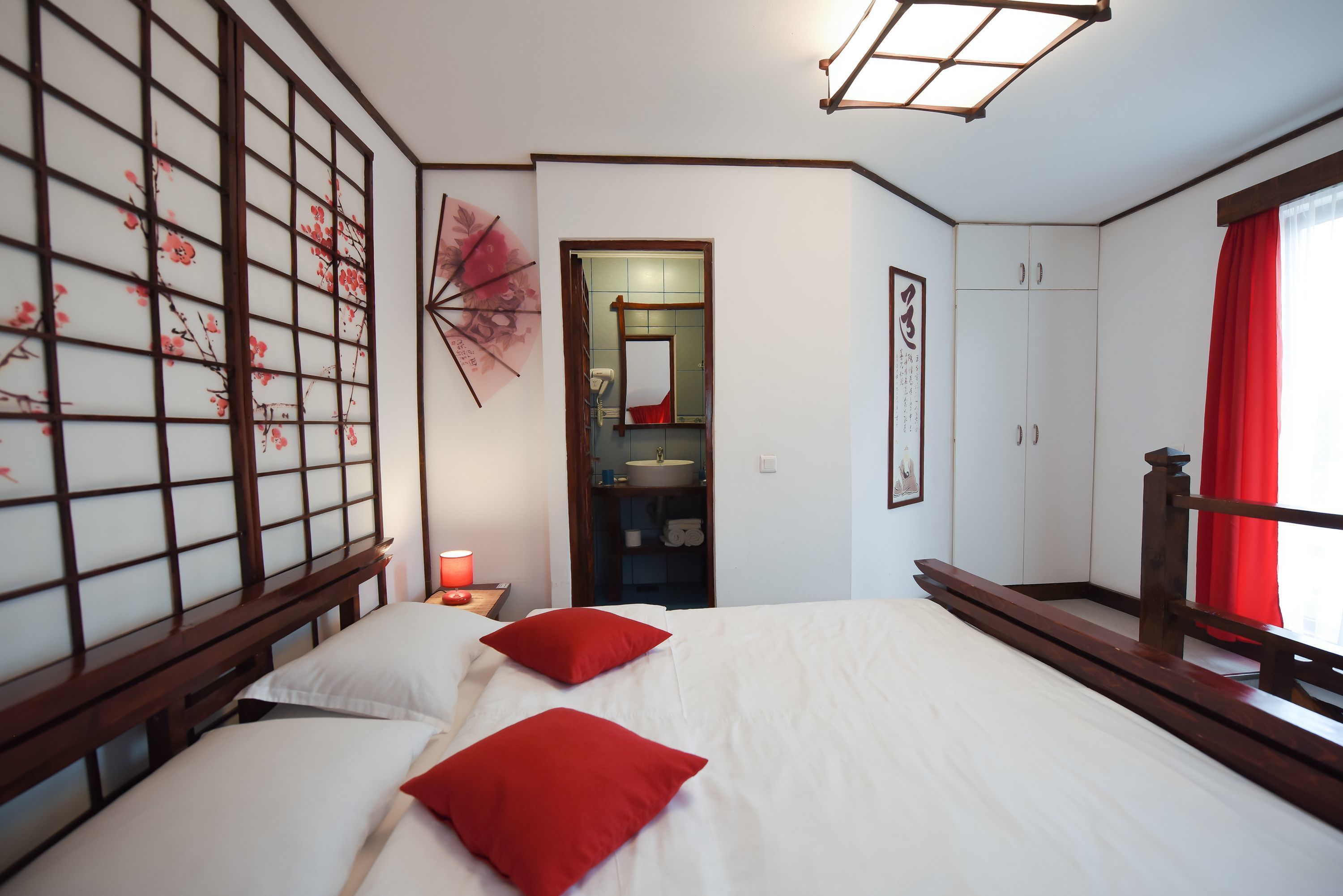 Ein Schlafzimmer mit einem Bett, roten Kissen, einem hölzernen Kopfteil, einem Nachttisch, einer roten Lampe, einem Spiegel, einem Kleiderschrank und einem Fenster mit roten Vorhängen.