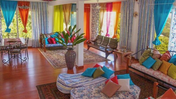 Un salon spacieux doté d’un plancher en bois, d’un ensemble de canapés, d’une table à manger avec des chaises et d’une grande fenêtre agrémentée de rideaux colorés.