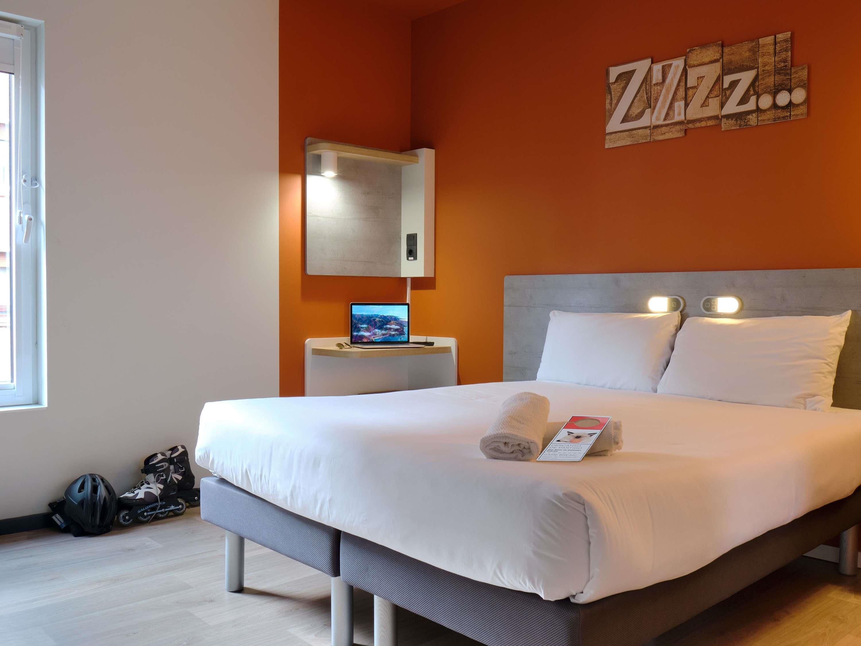 Una habitación de hotel moderna con una cama grande, una mesita de noche, una computadora portátil y un espejo en la pared.