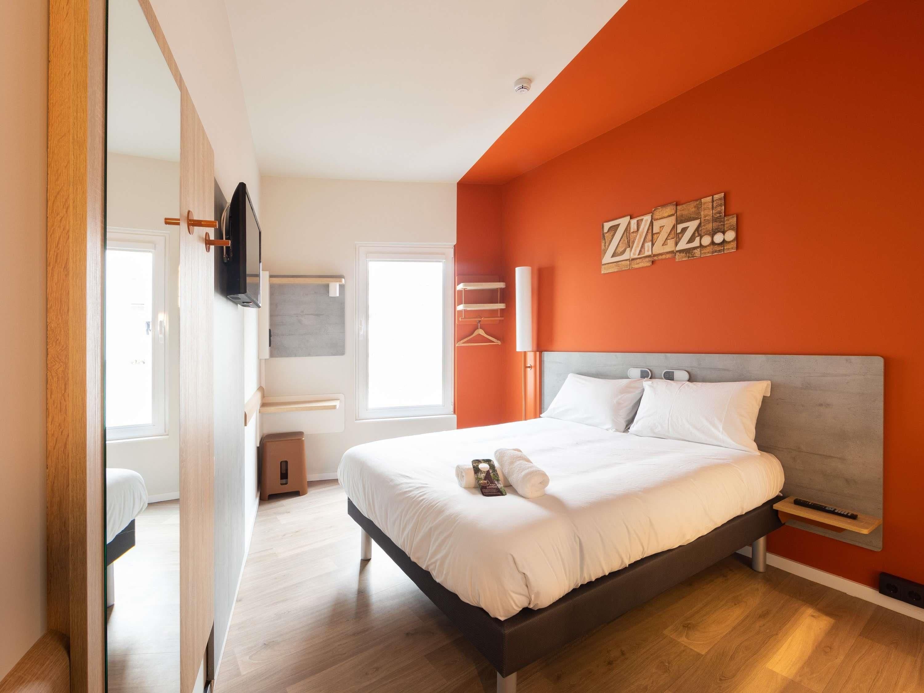 A modern bedroom with a large bed, a TV mounted on the wall, and a bright orange accent wall.