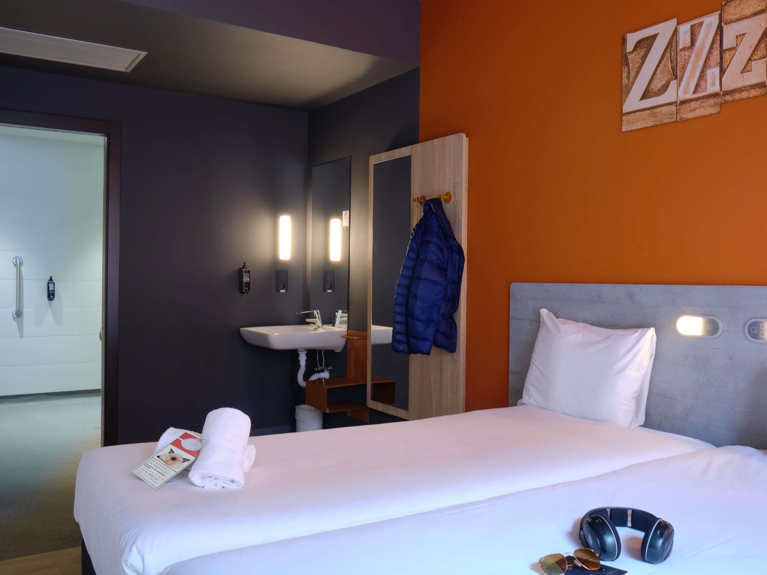 A hotel room with a bed, a bedside table, a mirror, a sink, and a coat rack.