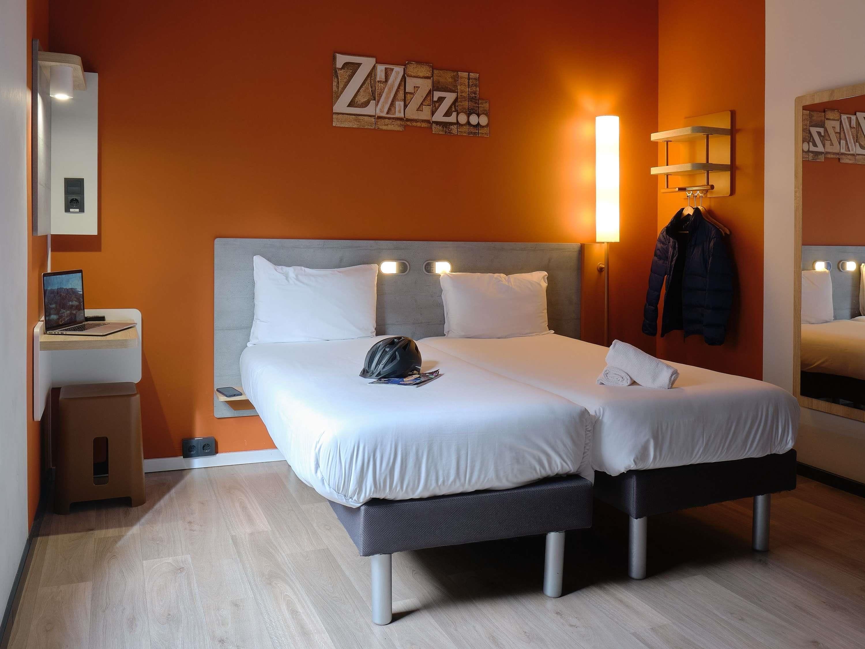 A double bed with a grey headboard, two white pillows, a black backpack, and a rolled towel on the bed.