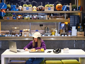 Una persona sentada a una mesa con una computadora portátil, un teléfono inteligente y una comida. La mesa es blanca, y la persona lleva un suéter púrpura y una gorra blanca.