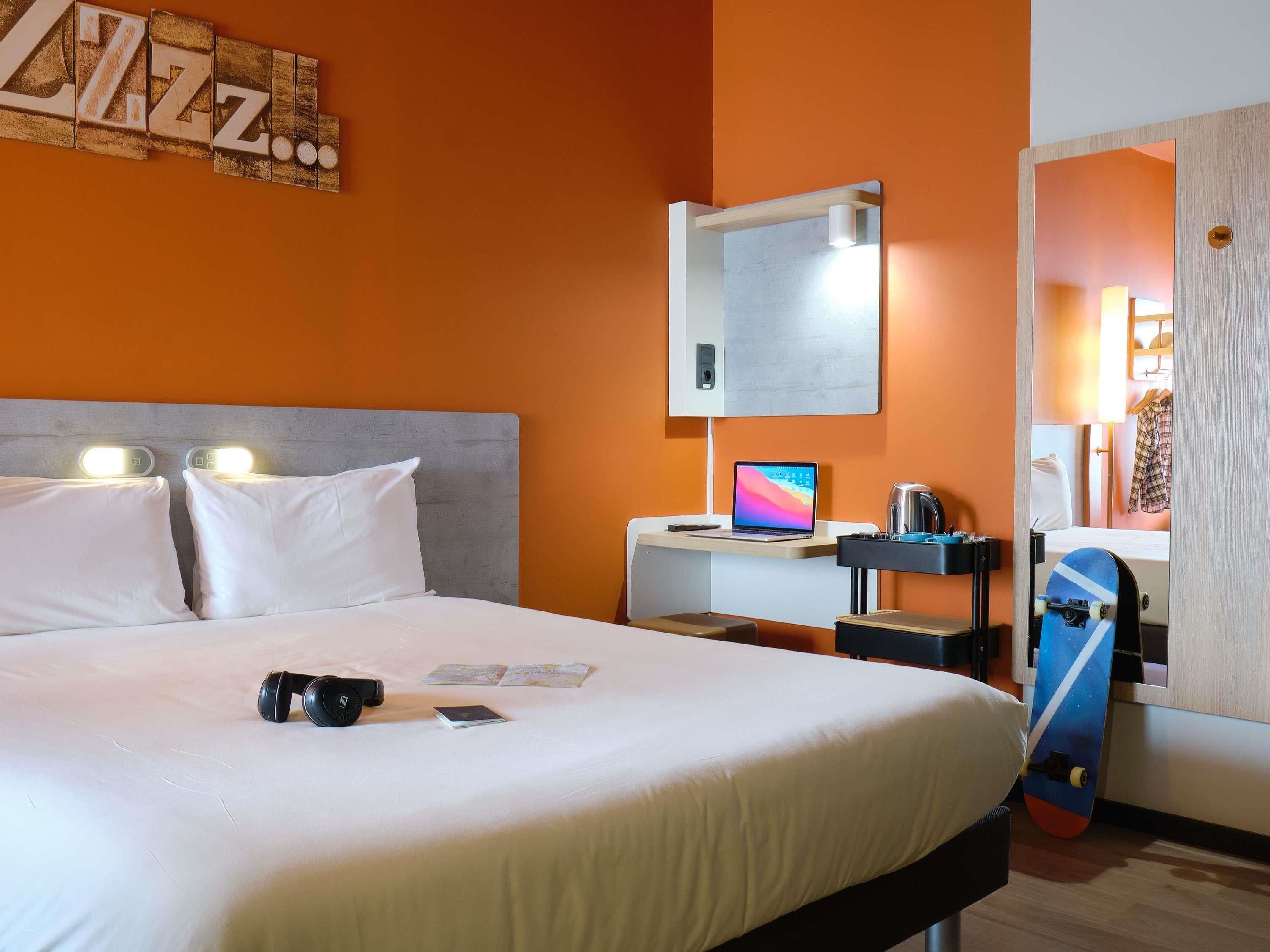 A hotel room with a bed, a desk with a laptop, a mirror, and a skateboard.