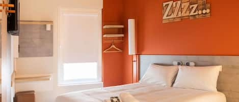 Una cama bien hecha con almohadas, una mesita de noche con control remoto, un televisor montado en la pared y un cartel con la leyenda 'Zzz'.