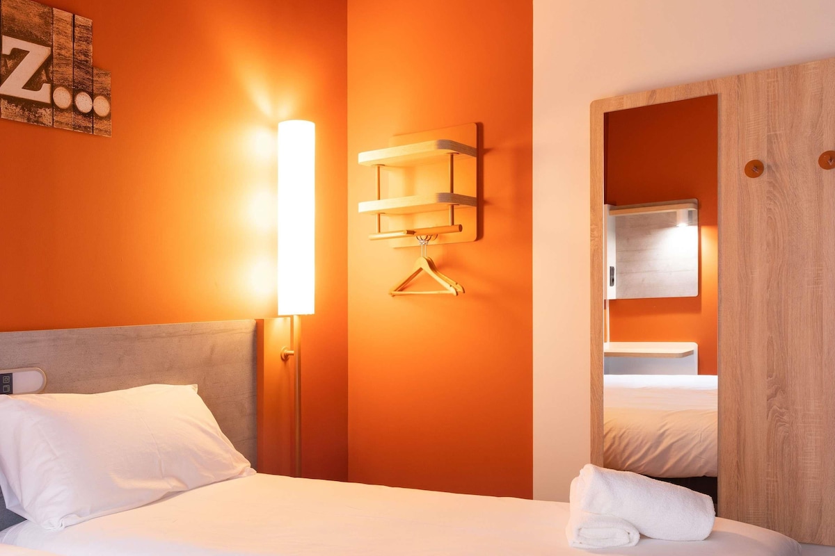 Una habitación de hotel con cama, lámpara de noche, armario de madera y aplique en la pared.