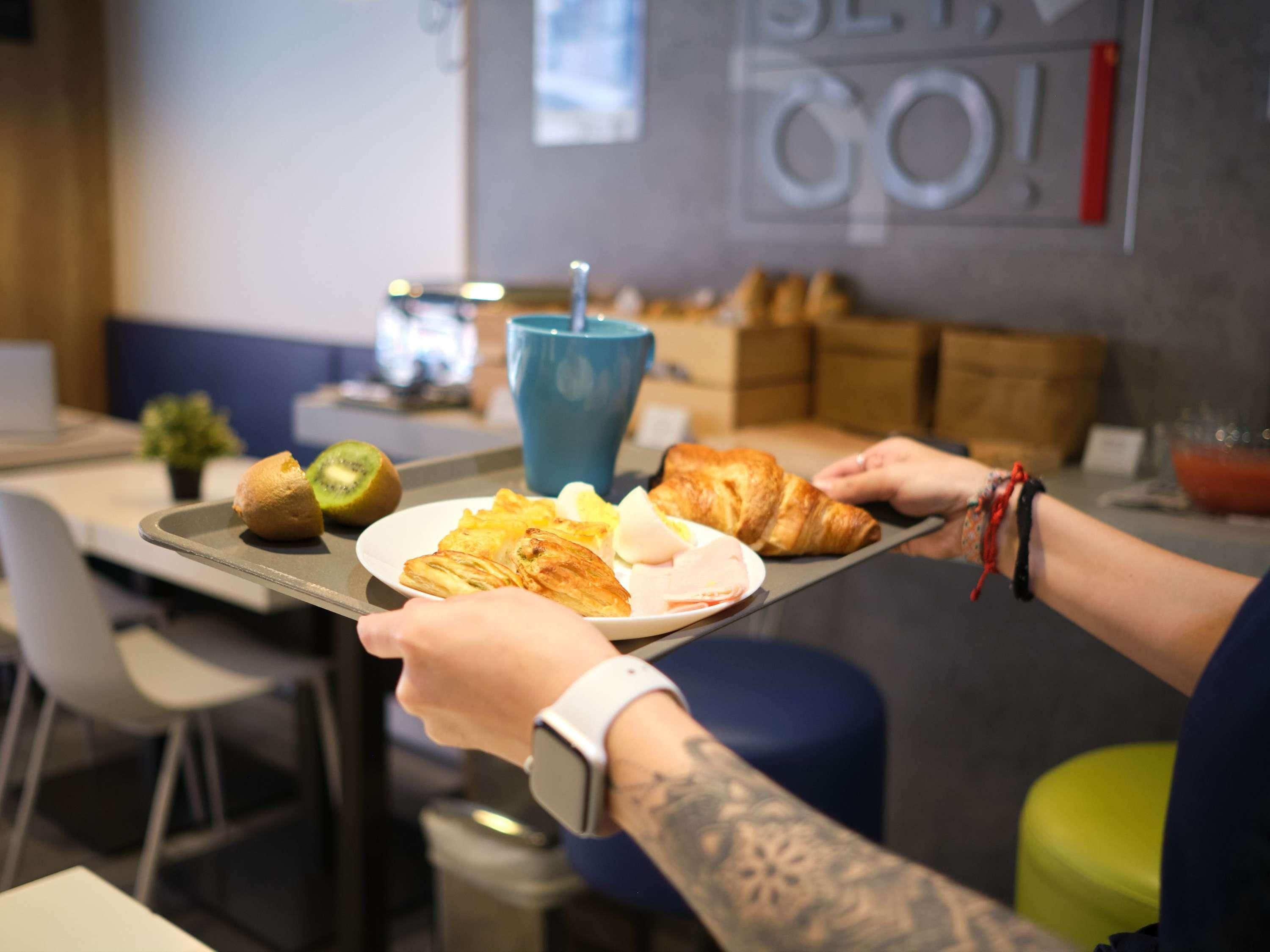 Un plateau contenant divers aliments, notamment des croissants, des œufs et un kiwi, tenu par une personne portant une montre connectée.