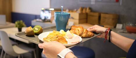 Un plateau contenant divers aliments, notamment des croissants, des œufs et un kiwi, tenu par une personne portant une montre connectée.