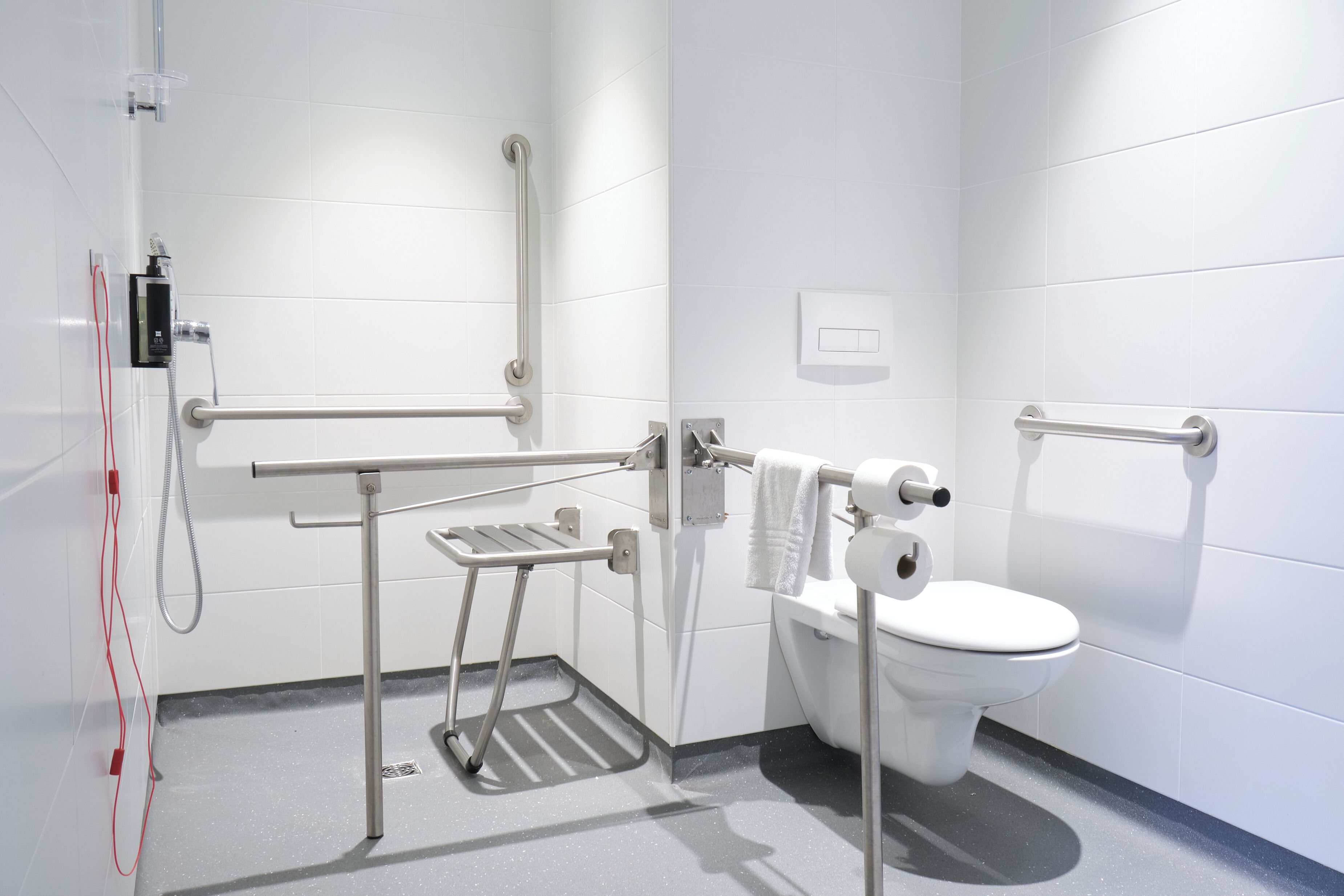 A modern accessible bathroom with a white toilet, grab bars, a shower with a handheld showerhead, and a hand dryer.