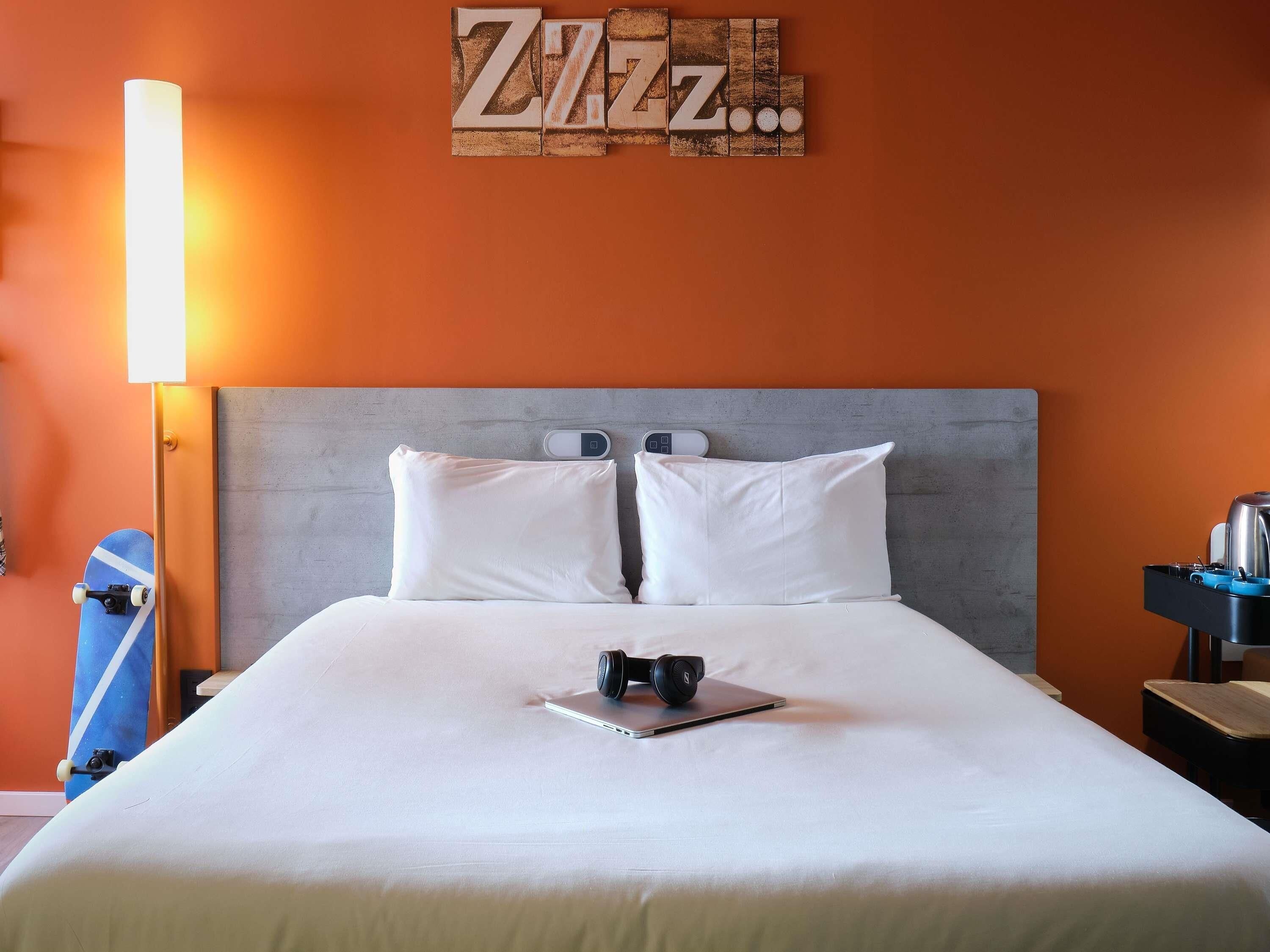 A hotel room with a bed, pillows, a bedside table with a kettle, and a wall with a 'ZAZZ...' sign.