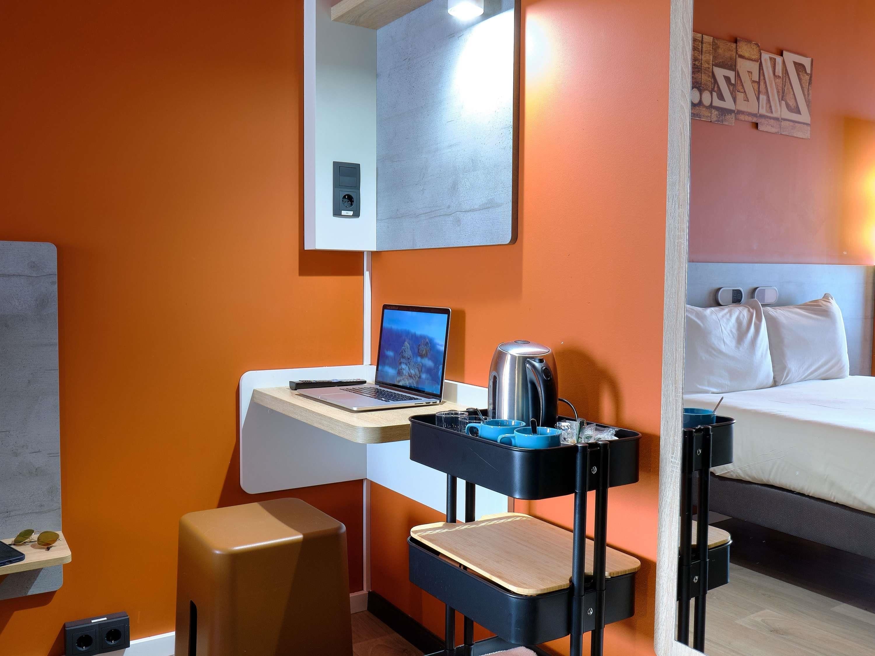 A compact hotel room with a bed, a desk with a laptop, a coffee maker, and a small seating area.
