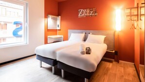 A hotel room with a bed, bedside tables, a mirror, a wall clock, and a window with a view of buildings.