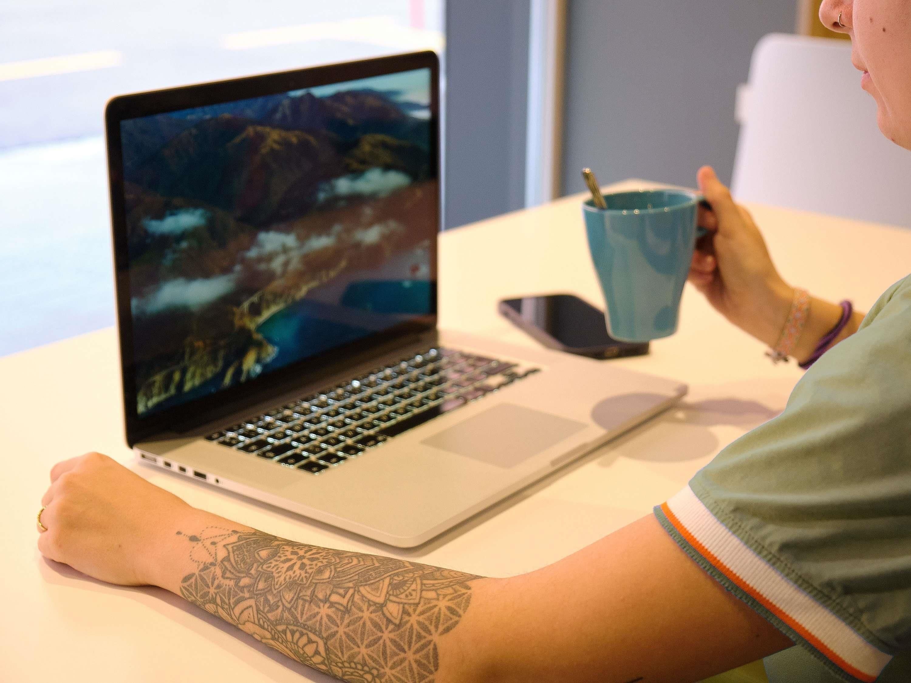 Une personne utilise un ordinateur portable dont l’écran affiche un paysage de montagne. Elle est assise à une table, avec un téléphone intelligent et une tasse bleue à portée de main.