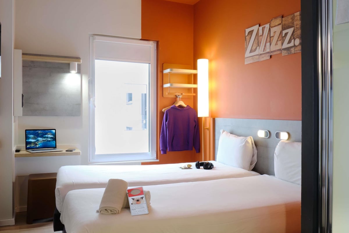Habitación de hotel con dos camas, un escritorio con una computadora portátil, una bata colgada y una lámpara fijada a la pared.