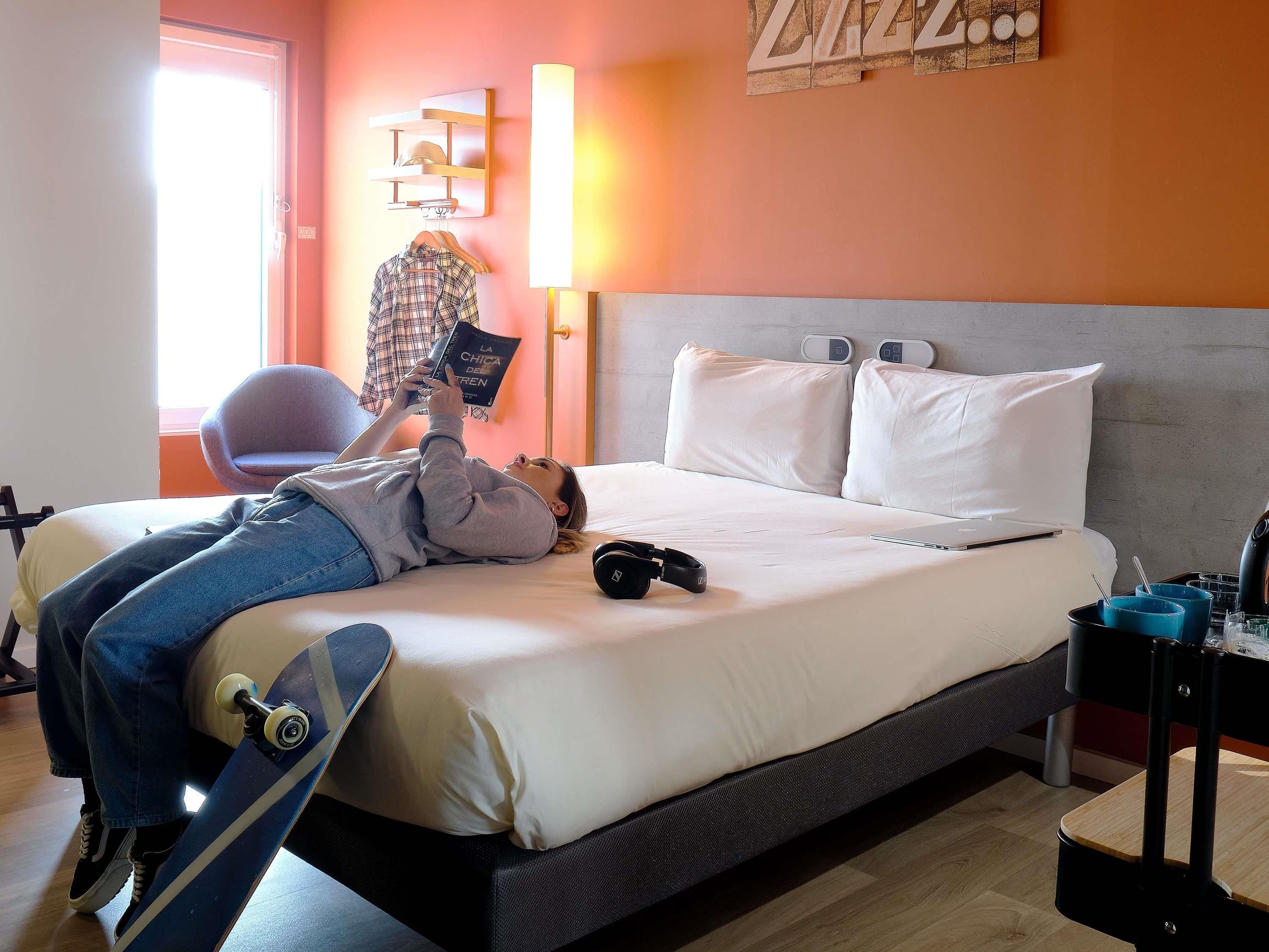 A hotel room with a bed, a chair, a desk with a laptop, and a skateboard.