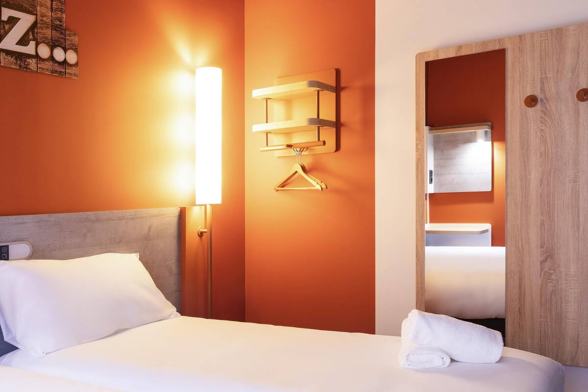 Una habitación de hotel con una cama, una lámpara de noche, un armario de madera y un perchero en la pared.