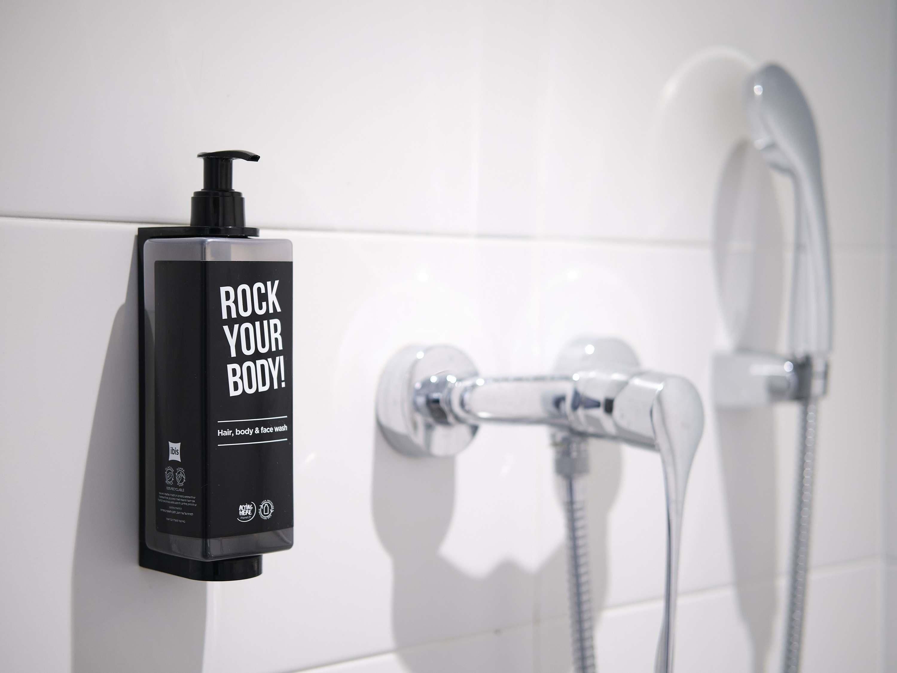 Una botella negra de gel de ducha con dosificador bomba, fijada a una pared de azulejos blancos, junto a una ducha con grifo y manguera cromados.